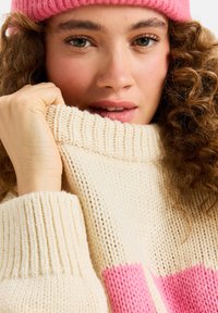 Gehaakte trui in crème met roze strepen, voorzien van een hoge geribbelde kraag. De textuur lijkt zacht en chunky, wat de warmte van het materiaal benadrukt.