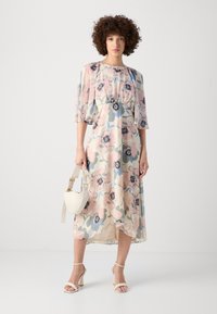 Floral midi dress in light pastel colors with puff sleeves, cinched waist, and flowing fabric. Paired with a cream handbag and strappy heels.
