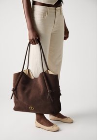 Sac fourre-tout en daim brun avec deux anses en cuir brun, deux nœuds à l'avant et un logo doré en bas. Le modèle porte un pantalon beige et des chaussures.