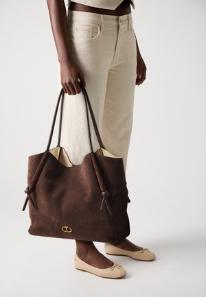 Borsa tote in suede marrone con due manici in pelle marrone, due nodi sul fronte e un logo dorato in basso. Il modello indossa pantaloni e scarpe beige.