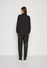 Black long-sleeve shirt with a collar, soft fabric, relaxed fit, back pleat details; paired with black leather pants and white sneakers.