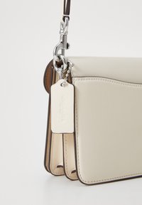 Cream leather bag with a structured design, silver hardware, and a tag displaying the brand name. Includes contrasting brown trim.