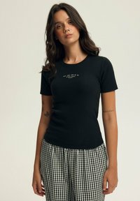 Czarny żeberkowy T-shirt z krótkimi rękawami i okrągłym dekoltem, z białym tekstem umieszczonym na środku klatki piersiowej. W połączeniu z czarno-białymi spodniami w kratę.