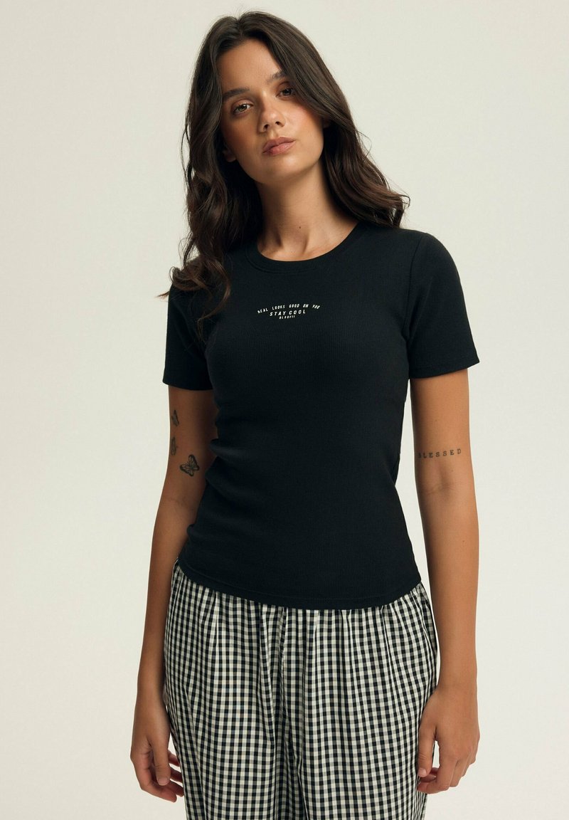 Czarny żeberkowy T-shirt z krótkimi rękawami i okrągłym dekoltem, z białym tekstem umieszczonym na środku klatki piersiowej. W połączeniu z czarno-białymi spodniami w kratę.