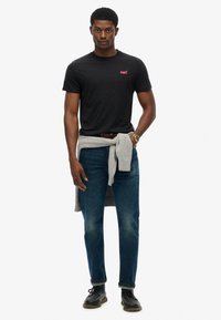 Zwarte crew neck t-shirt met roze logo, gecombineerd met lichtblauwe denim jeans en een grijze trui om de taille geknoopt. Zwarte schoenen maken de look af.