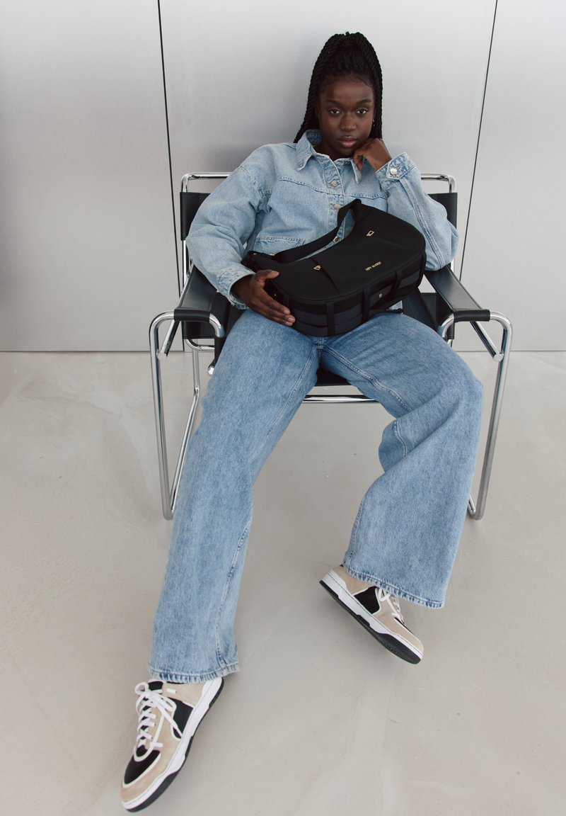 Zwarte schoudertas met gouden accenten, gecombineerd met een lichtblauwe spijkerjacket, losse blauwe spijkerbroek en zwart-witte sneakers.