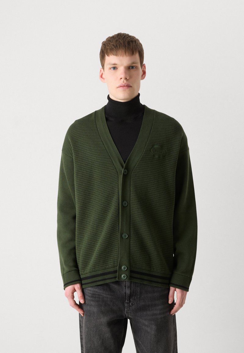 Zielony kardigan z dzianiny z dekoltem w kształcie litery V, z zapięciem na guziki i teksturalnym wzorem. Noszony na czarnym golfie, w połączeniu z ciemnymi dżinsami.