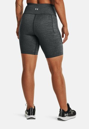 Athlète portant un short de sport moulant gris foncé et des baskets noires Under Armour, debout de dos sur un fond blanc.