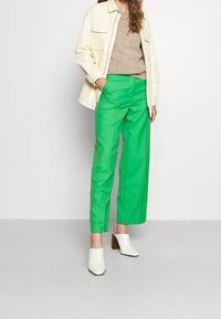 Vrouw in felgroene wijde broek, beige trui, crème jas en witte hakken op een effen achtergrond.