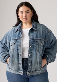 Light blue denim jacket with a relaxed fit, featuring two front pockets, metal buttons, and a classic collar. Worn over a white top.