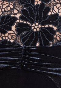 Zwart kanten lijfje met bloemenpatroon op naakte stof; geplooide zwarte fluwelen tailleband voegt textuur en contrast toe.