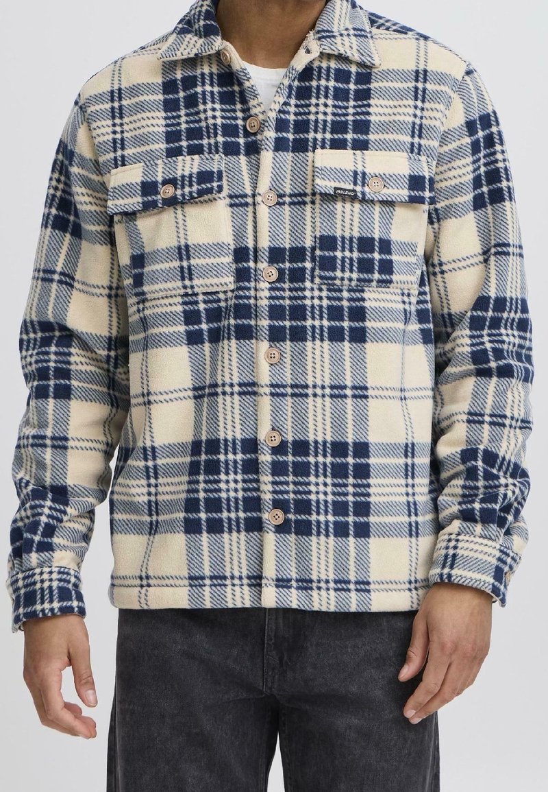 Homme portant une veste polaire à carreaux beige et bleu marine boutonnée avec deux poches poitrine et un pantalon noir.