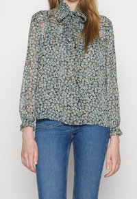 Blusa negra translúcida con un patrón floral en tonos azules y verdes, con un cuello de lazo y puños elásticos, combinada con vaqueros azules.