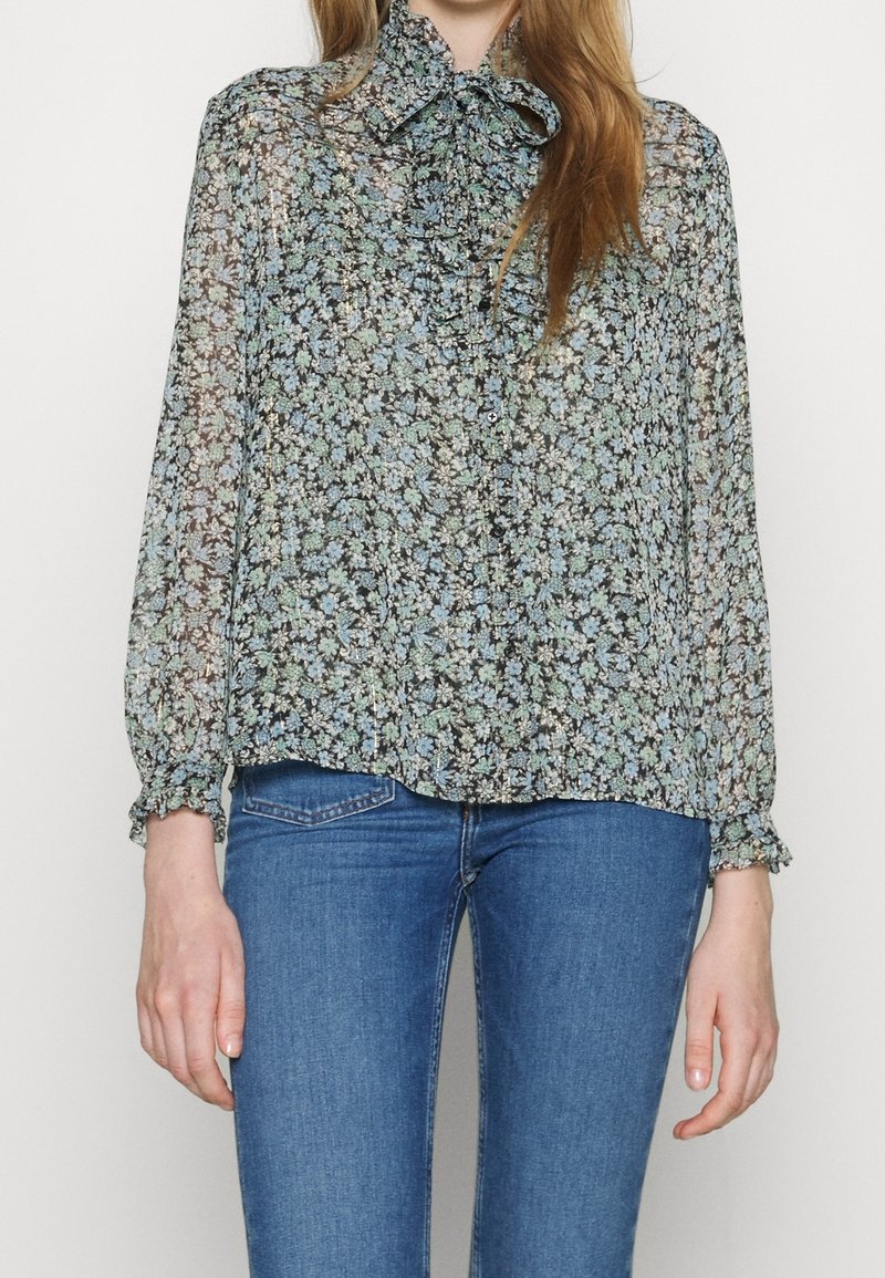 Blusa negra translúcida con un patrón floral en tonos azules y verdes, con un cuello de lazo y puños elásticos, combinada con vaqueros azules.