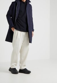 Marineblauer Mantel mit hohem Kragen und Tasche, schwarzes T-Shirt, cremefarbene plissierte Hose und schwarze Sneakers. Glatte Stoffe und lockere Passform.