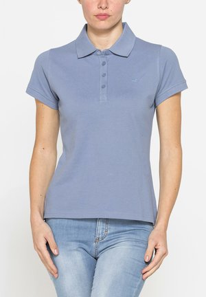 Donna che indossa una polo blu aderente a maniche corte con bottoni e jeans di denim azzurro chiaro in piedi davanti a uno sfondo semplice.