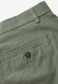 Olivgrüne Baumwollhosen mit einer Gesäßtasche, die mit einem schwarzen Knopf und tonal abgestimmten Nähten versehen ist. Glatte Textur und maßgeschneiderte Designdetails.