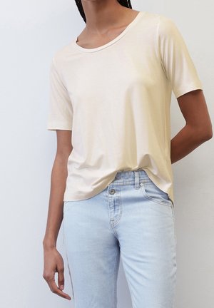 Person trägt ein schlichtes hellbeiges Kurzarm-T-Shirt, das leicht in hellblaue Jeans gesteckt ist, vor weißem Hintergrund.