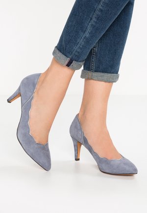 Vrouw die lichtblauwe suède pumps met puntige neus, gegolfde randen en omgeslagen donkerblauwe spijkerbroek draagt, tegen een witte achtergrond.