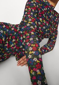 Person, der sich zurücklehnt und ein langärmeliges, eng anliegendes Kleid mit einem bunten Blumenmuster auf schwarzem Hintergrund trägt.