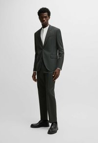 Abito grigio scuro a righe con rever a taglio, pantaloni coordinati e una camicia bianca sotto. Scarpe eleganti nere completano il look.