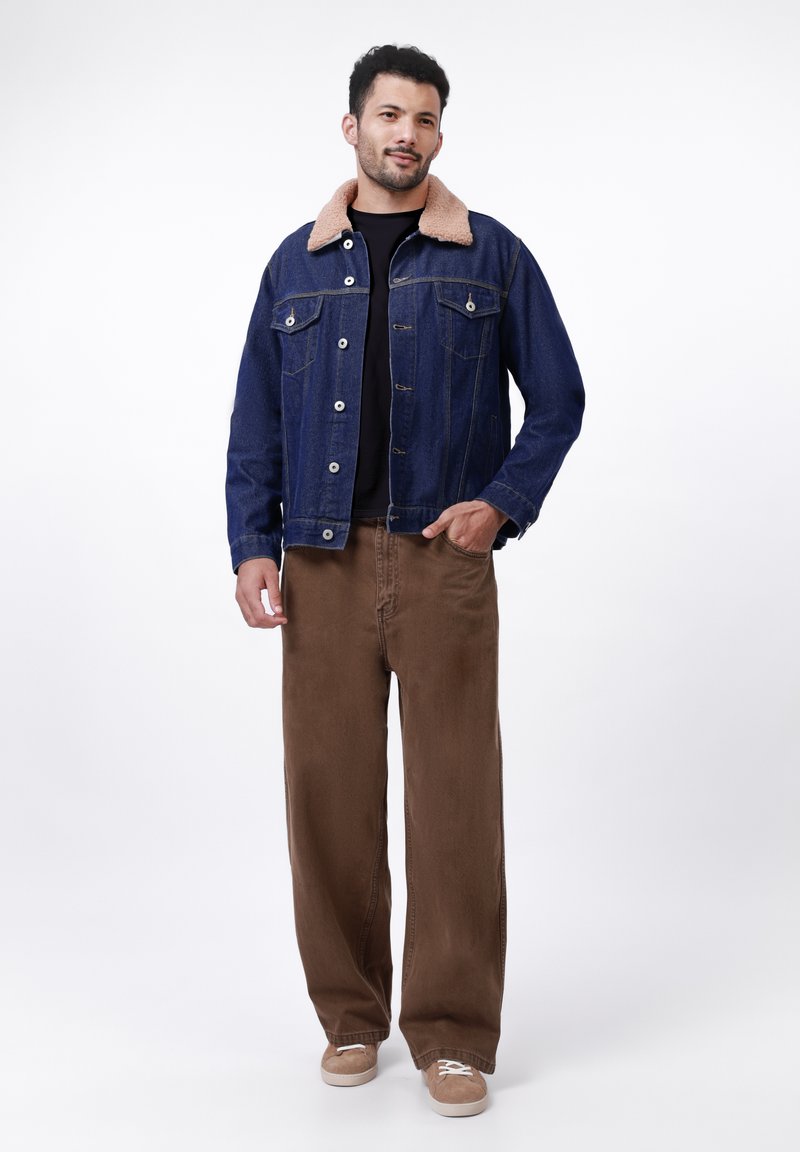 Veste en denim avec un col en fausse fourrure beige, tissu bleu, poches avant, associée à un pantalon large marron et des baskets beiges.