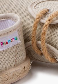 Chaussure en fibres naturelles tressées de couleur beige avec une texture tissée. Présente une étiquette colorée cousue et une semelle en jute avec un détail de laçage.