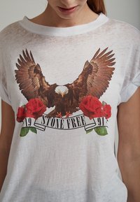 Hvid kortærmet t-shirt med en brun ørn-grafik over røde roser og et banner med teksten "19 STONE FREE 91." Blødt, let gennemsigtigt stof.