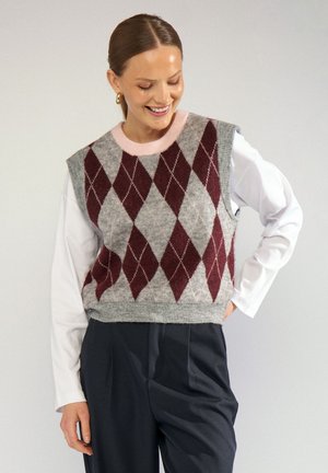 Femme portant un gilet en maille gris et bordeaux à motif losanges par-dessus une chemise blanche à manches longues avec un pantalon plissé foncé, souriant et regardant vers le bas.