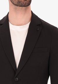 Blazer nero con rever a dente, dettaglio a un bottone e taschino sul petto. Indossato sopra una t-shirt bianca semplice. Tessuto dalla superficie liscia.