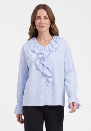 Woman wearing light blue blouse with ruffled eyelet details on collar and cuffs, paired with black pants, standing against white background.