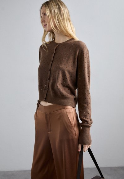 Καφέ πλεκτό ζακέτα με μικρές υφασμένες διακοσμήσεις, μακριά μανίκια και μπροστινό κουμπί, συνδυασμένη με σατέν καφέ πα�ντελόνια σε φαρδύ ίσιο κόψιμο.