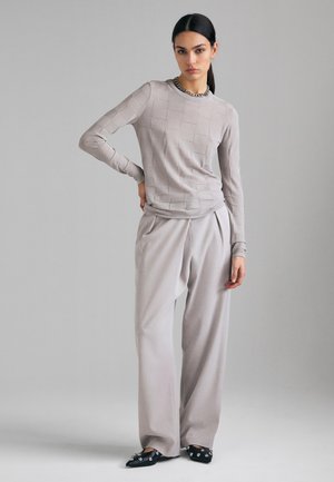 Donna con capelli scuri che indossa un maglione grigio chiaro con trama, pantaloni larghi grigio pallido, ballerine nere con punta appuntita e decorazioni, e una collana a catena color argento.