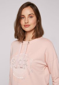 Felpa con cappuccio rosa chiaro con design grafico frontale che presenta testi e motivi a pois variabili. Include stringa di regolazione e accenti metallici.