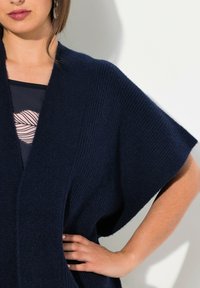 Marinblå stickad kofta med vida ärmar, ribbad textur och öppen front. Lättviktsmaterial, mjukt, med en avslappnad silhuett.