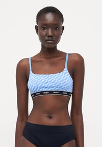 Light blue bralette with white "HUGO" logo pattern, narrow straps, and a black elastic band featuring the logo. Smooth fabric texture.