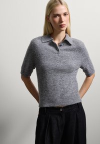 Grauer kurzärmeliger Polopullover aus weichem Strickmaterial mit Kragen und geripptem Saum; getragen mit dunklen, gefalteten Hosen.