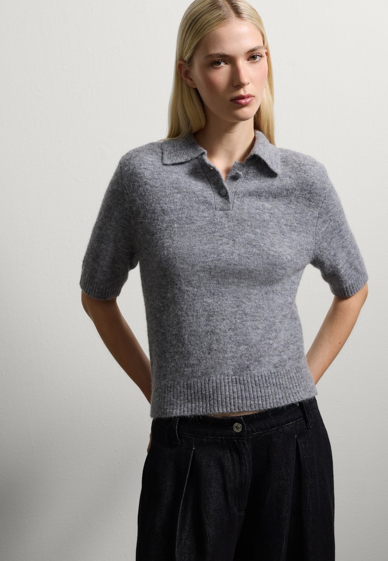 Grauer kurzärmeliger Polopullover aus weichem Strickmaterial mit Kragen und geripptem Saum; getragen mit dunklen, gefalteten Hosen.