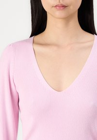 Top canelado em rosa suave com decote em V. Apresenta mangas longas com canelado ajustado e costura detalhada ao longo do decote.