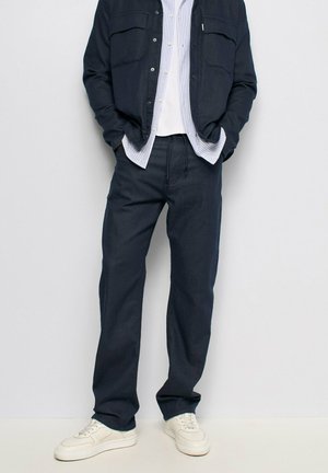 Homme portant un pantalon bleu foncé, des baskets blanches, un t-shirt blanc, une chemise rayée et une veste bleu foncé avec les mains dans les poches.