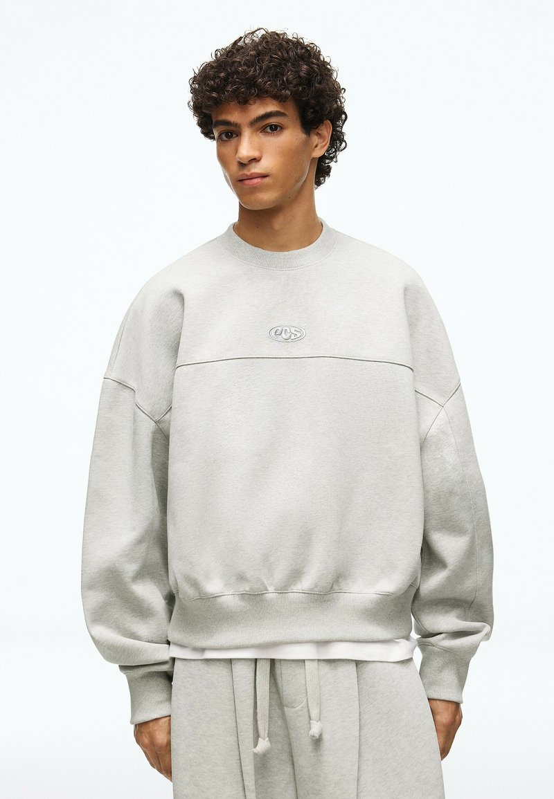 Jeune homme aux cheveux bouclés portant un sweat-shirt oversize gris clair et un pantalon de survêtement assorti sur fond blanc uni.
