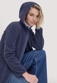 Marine fleece jack met capuchon, met een volle rits en een textuurstof. Gecombineerd met lichtblauwe denim, zittende pose.