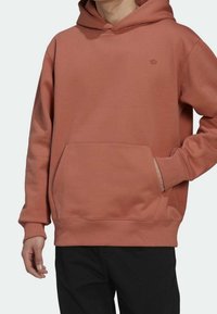 Man som bär en rostfärgad hoodie med framficka och liten logotyp, i kombination med svarta byxor, där handen vilar i fickan.