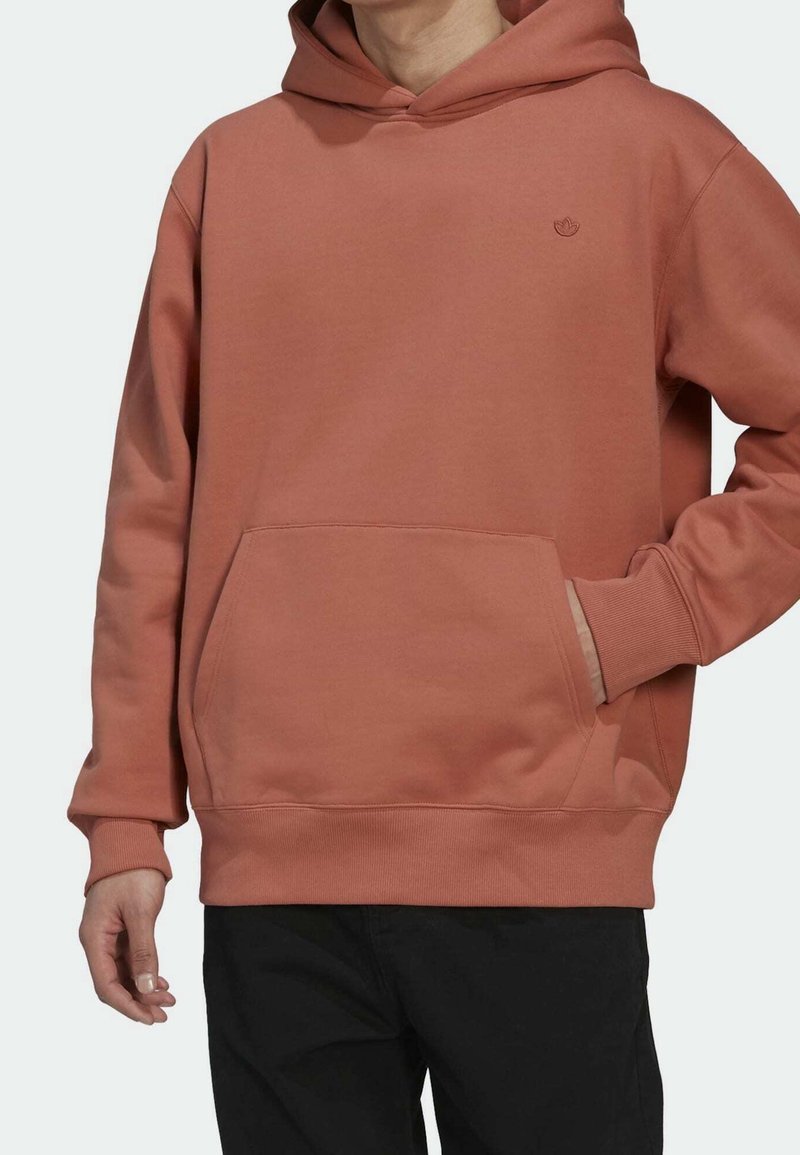 Man som bär en rostfärgad hoodie med framficka och liten logotyp, i kombination med svarta byxor, där handen vilar i fickan.