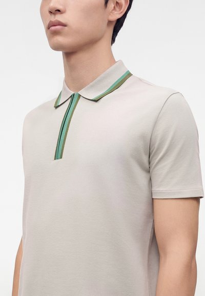 Homme portant un polo beige clair à manches courtes avec des rayures vertes et bleues sur le col et la patte de boutonnage, visible des épaules jusqu'au milieu du torse.