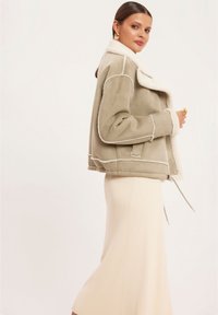 Jasnozielona kurtka z owczej wełny z miękkim, białym kołnierzem z futra, kontrastowymi przeszyciami i kieszeniami bocznymi, zestawiona z kremową spódnicą o prążkowanej fakturze.