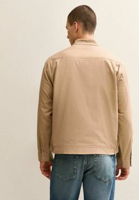 Uomo con giacca beige e jeans blu, in piedi di spalle contro uno sfondo chiaro e semplice.