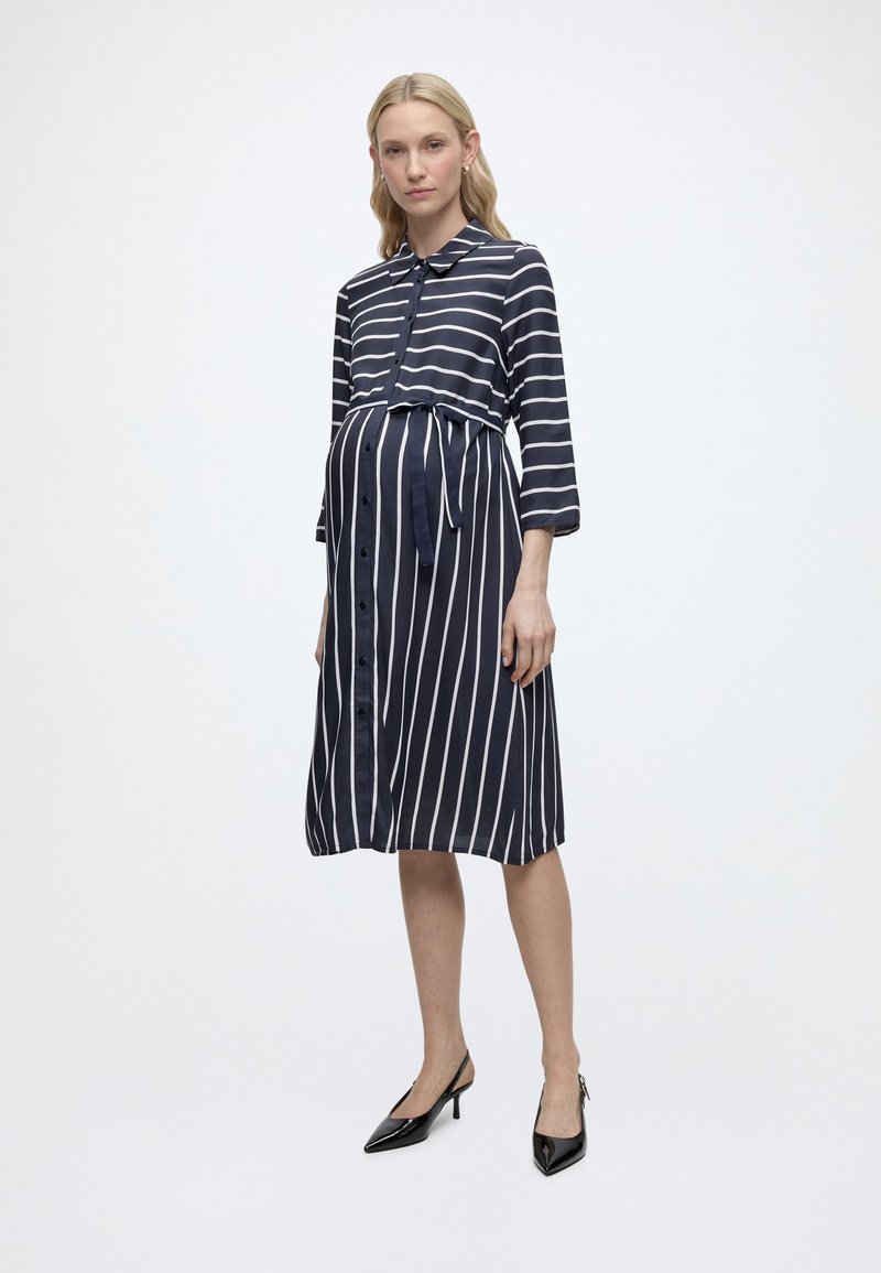 Έγκυος γυναίκα ντυμένη με μαύρο και άσπρο ριγέ φόρεμα εγκυμοσύνης με κουμπιά και ζώνη, στέκεται σε μαύρα τακούνια σε απλό φόντο.