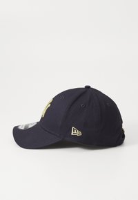 Navy blue baseball cap with a curved visor, featuring a gold embroidered logo on the front and a small tag on the side. Cotton material.