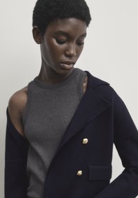 Granatowy żakiet z mieszanki wełny z złotymi guzikami, założony na szary bezrękawnik o gładkiej fakturze i dopasowanym kroju.